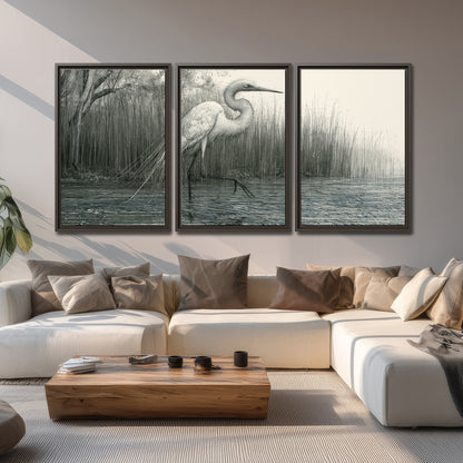 A pale long-legged heron standing beside tall reeds in a muted gray-green wetland 3 piece framed canvas wall art displayed in a living room