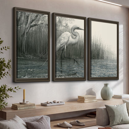 A pale long-legged heron standing beside tall reeds in a muted gray-green wetland 3 piece framed canvas wall art displayed in a console