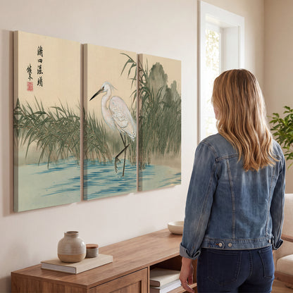 A white heron standing beside a quiet river with reeds and distant hills 3 piece canvas wall art three panel arrangement on a wall