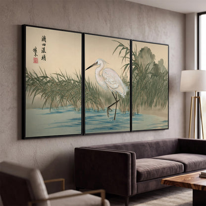 A white heron standing beside a quiet river with reeds and distant hills 3 piece framed canvas wall art displayed in a modern living room