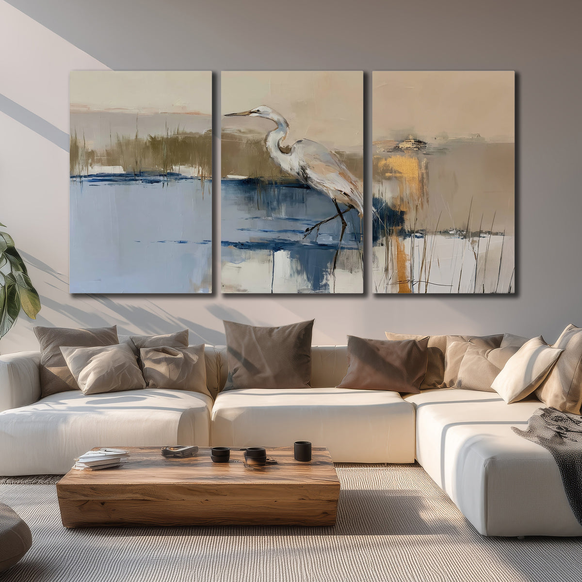 A slender heron standing in blue water among loose ochre and cream brushstrokes 3 piece canvas wall art displayed in a living room