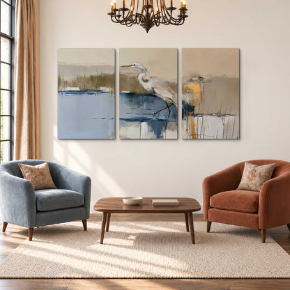 A slender heron standing in blue water among loose ochre and cream brushstrokes 3 piece canvas wall art styled in an interior