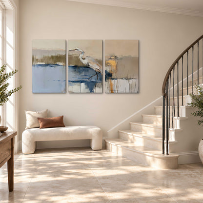 A slender heron standing in blue water among loose ochre and cream brushstrokes 3 piece canvas wall art styled in an interior