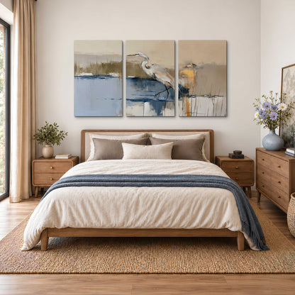A slender heron standing in blue water among loose ochre and cream brushstrokes 3 piece canvas wall art styled in an interior