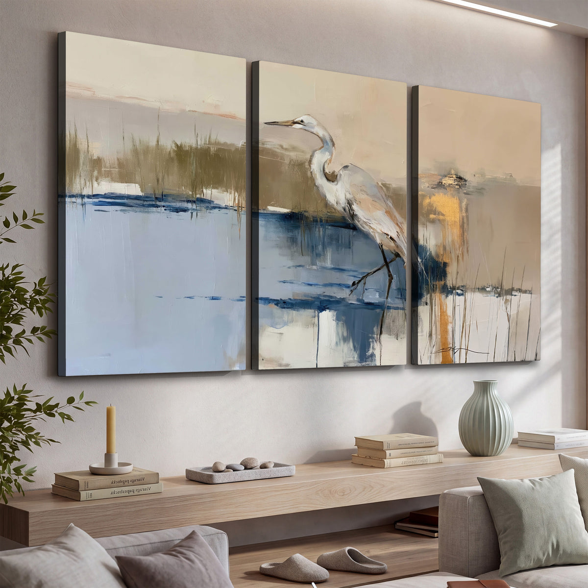 A slender heron standing in blue water among loose ochre and cream brushstrokes 3 piece canvas wall art displayed in a console