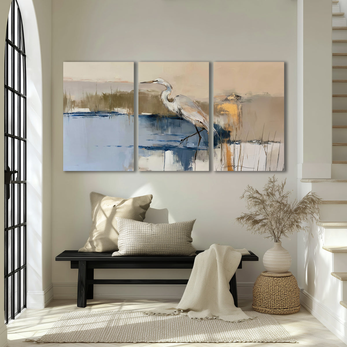 A slender heron standing in blue water among loose ochre and cream brushstrokes 3 piece canvas wall art displayed in a entryway