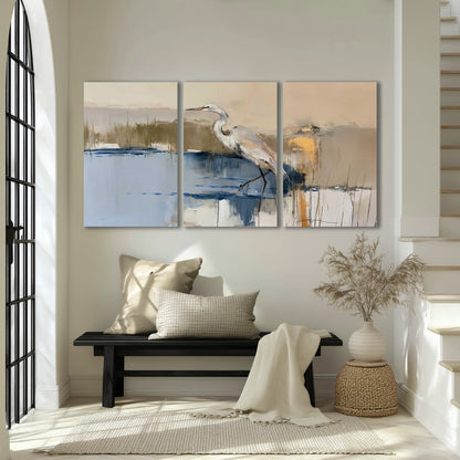 A slender heron standing in blue water among loose ochre and cream brushstrokes 3 piece canvas wall art displayed in a entryway