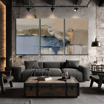 A slender heron standing in blue water among loose ochre and cream brushstrokes 3 piece canvas wall art displayed in a industrial room