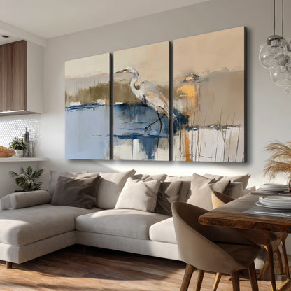 A slender heron standing in blue water among loose ochre and cream brushstrokes 3 piece canvas wall art room and product view
