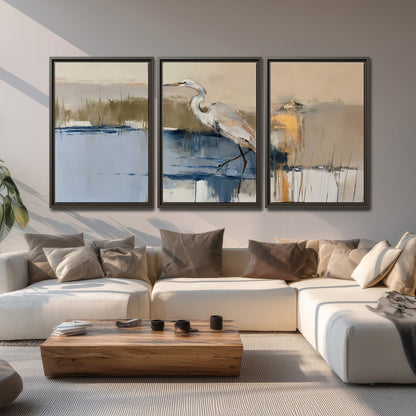 A slender heron standing in blue water among loose ochre and cream brushstrokes 3 piece framed canvas wall art displayed in a living room