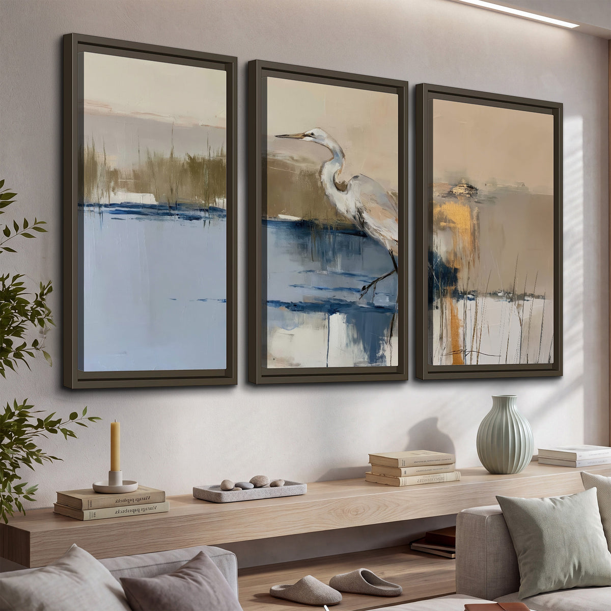 A slender heron standing in blue water among loose ochre and cream brushstrokes 3 piece framed canvas wall art displayed in a console