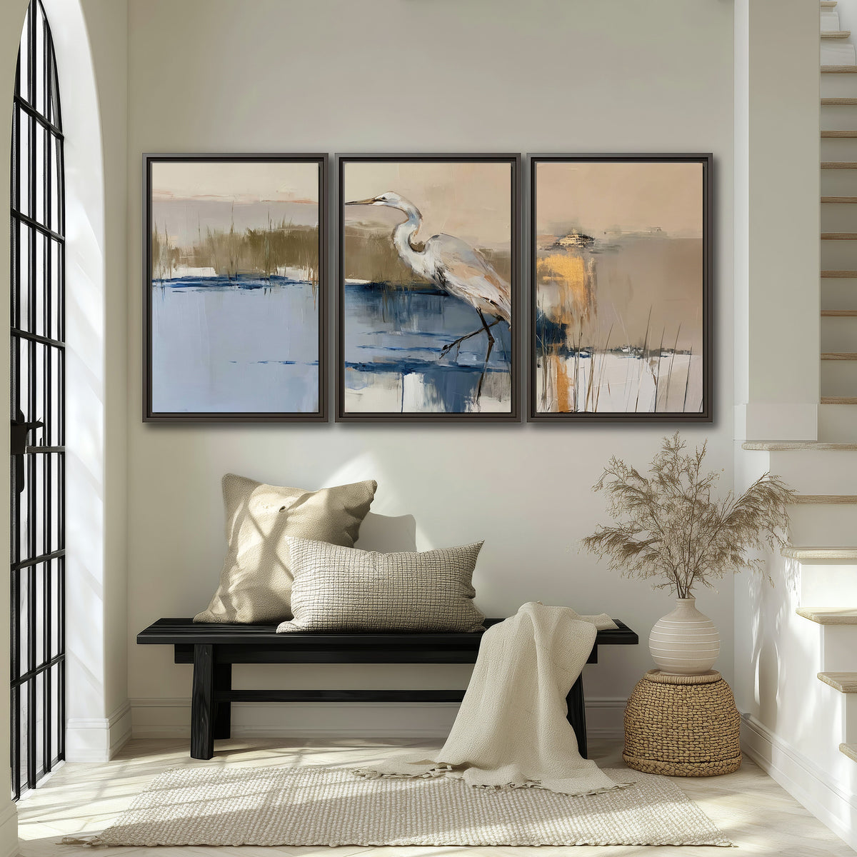 A slender heron standing in blue water among loose ochre and cream brushstrokes 3 piece framed canvas wall art displayed in a entryway