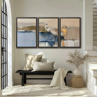 A slender heron standing in blue water among loose ochre and cream brushstrokes 3 piece framed canvas wall art displayed in a entryway