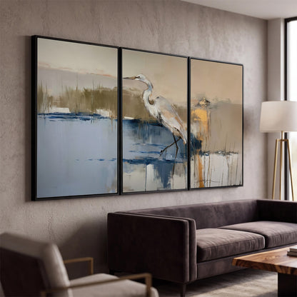 A slender heron standing in blue water among loose ochre and cream brushstrokes 3 piece framed canvas wall art displayed in a modern living room