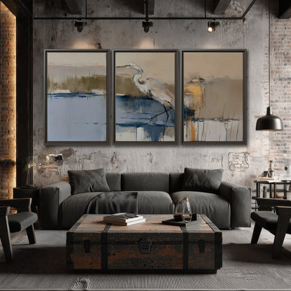 A slender heron standing in blue water among loose ochre and cream brushstrokes 3 piece framed canvas wall art displayed in a industrial room