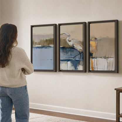 A slender heron standing in blue water among loose ochre and cream brushstrokes 3 piece framed canvas wall art three panel arrangement on a wall