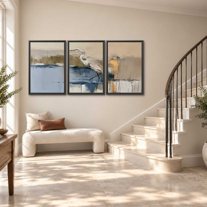 A slender heron standing in blue water among loose ochre and cream brushstrokes 3 piece framed canvas wall art styled in an interior