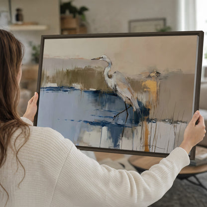 A slender heron standing in blue water among loose ochre and cream brushstrokes single framed canvas wall art held in hands