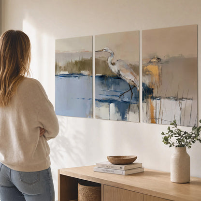 A slender heron standing in blue water among loose ochre and cream brushstrokes set of 3 poster prints three panel arrangement on a wall