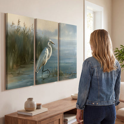 A white heron beside tall marsh grasses and still blue water 3 piece canvas wall art three panel arrangement on a wall