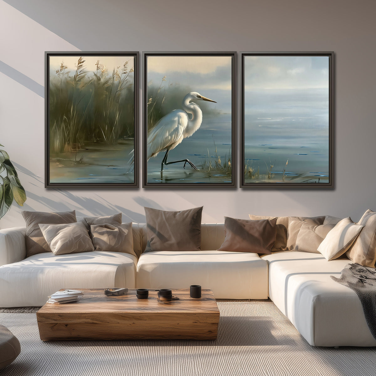 A white heron beside tall marsh grasses and still blue water 3 piece framed canvas wall art displayed in a living room