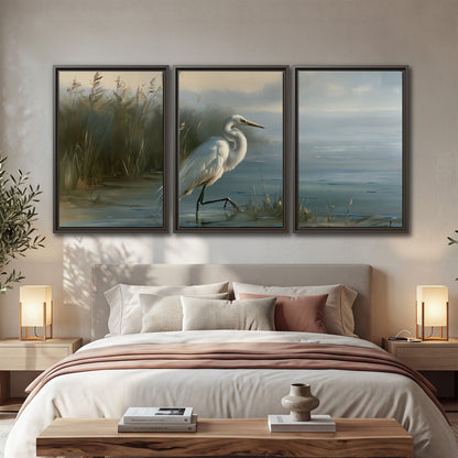 A white heron beside tall marsh grasses and still blue water 3 piece framed canvas wall art displayed in a bedroom