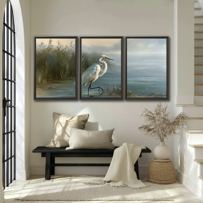 A white heron beside tall marsh grasses and still blue water 3 piece framed canvas wall art displayed in a entryway
