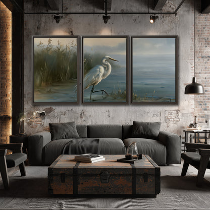 A white heron beside tall marsh grasses and still blue water 3 piece framed canvas wall art displayed in a industrial room