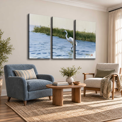 A white heron wading through bright blue water beside green reeds 3 piece canvas wall art styled in an interior