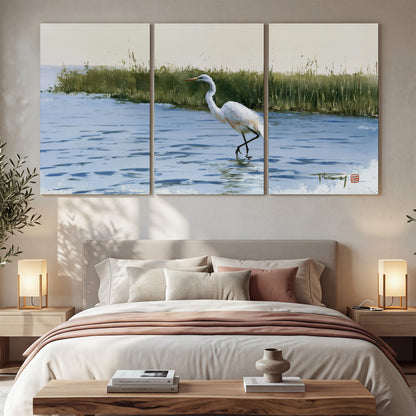 A white heron wading through bright blue water beside green reeds 3 piece canvas wall art displayed in a bedroom