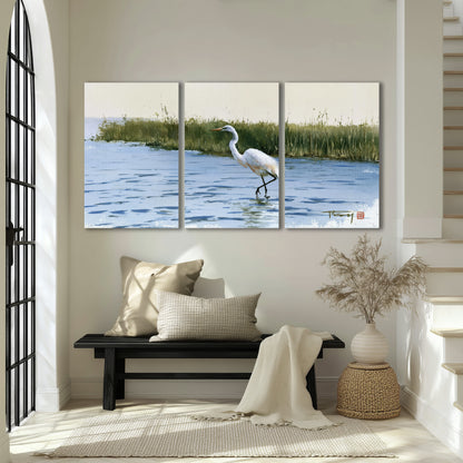 A white heron wading through bright blue water beside green reeds 3 piece canvas wall art displayed in a entryway