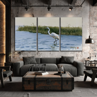 A white heron wading through bright blue water beside green reeds 3 piece canvas wall art displayed in a industrial room
