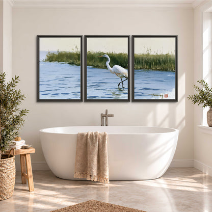 A white heron wading through bright blue water beside green reeds 3 piece framed canvas wall art styled in an interior