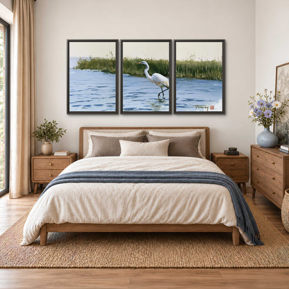 A white heron wading through bright blue water beside green reeds 3 piece framed canvas wall art styled in an interior