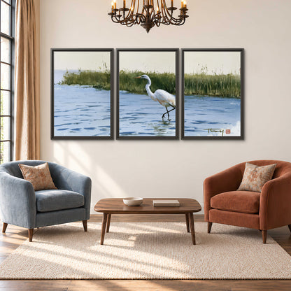 A white heron wading through bright blue water beside green reeds 3 piece framed canvas wall art styled in an interior