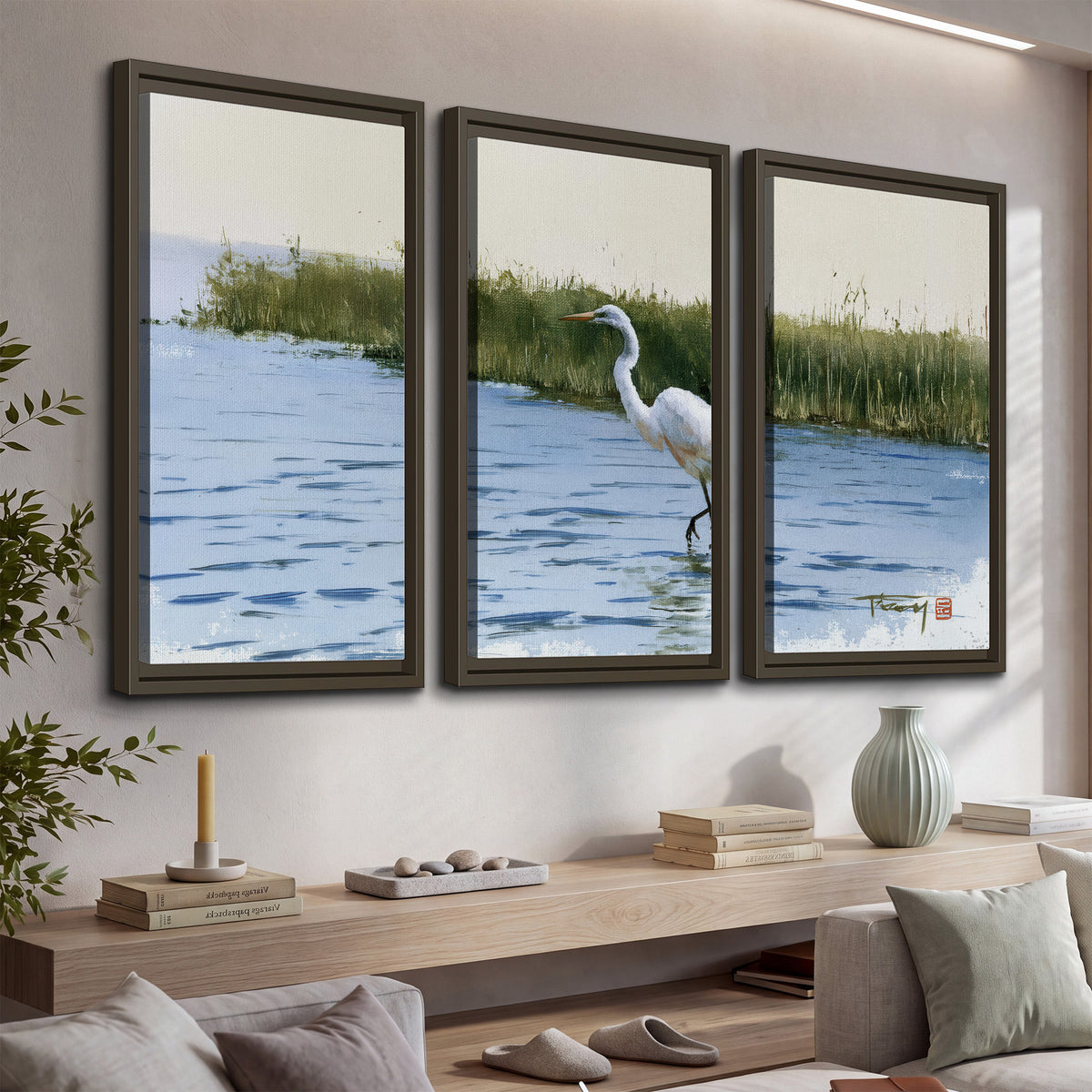 A white heron wading through bright blue water beside green reeds 3 piece framed canvas wall art displayed in a console