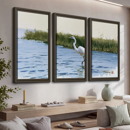 A white heron wading through bright blue water beside green reeds 3 piece framed canvas wall art displayed in a console