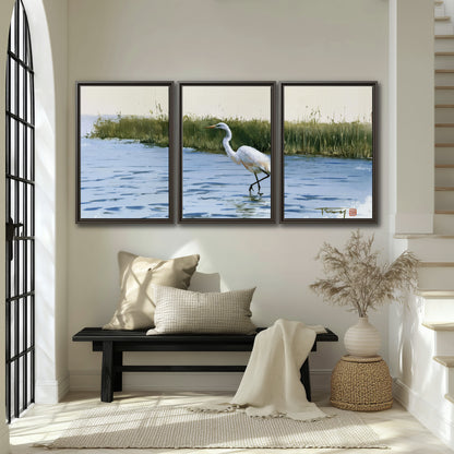A white heron wading through bright blue water beside green reeds 3 piece framed canvas wall art displayed in a entryway