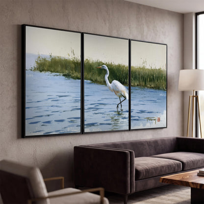 A white heron wading through bright blue water beside green reeds 3 piece framed canvas wall art displayed in a modern living room