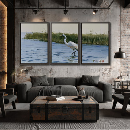 A white heron wading through bright blue water beside green reeds 3 piece framed canvas wall art displayed in a industrial room