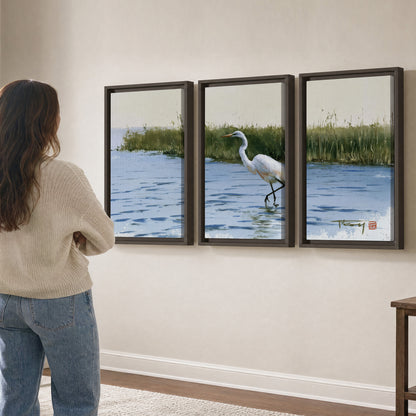 A white heron wading through bright blue water beside green reeds 3 piece framed canvas wall art three panel arrangement on a wall
