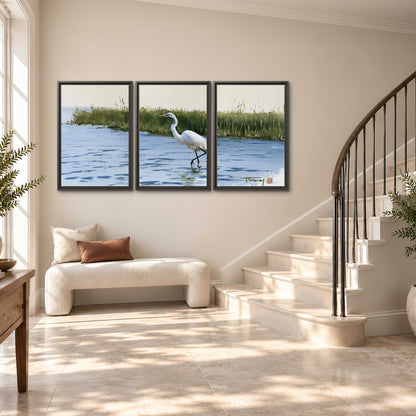 A white heron wading through bright blue water beside green reeds 3 piece framed canvas wall art styled in an interior