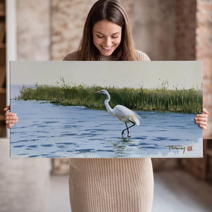 A white heron wading through bright blue water beside green reeds single canvas wall art held in hands