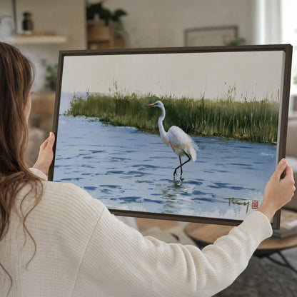 A white heron wading through bright blue water beside green reeds single framed canvas wall art held in hands
