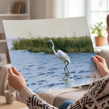 A white heron wading through bright blue water beside green reeds single poster print held in hands