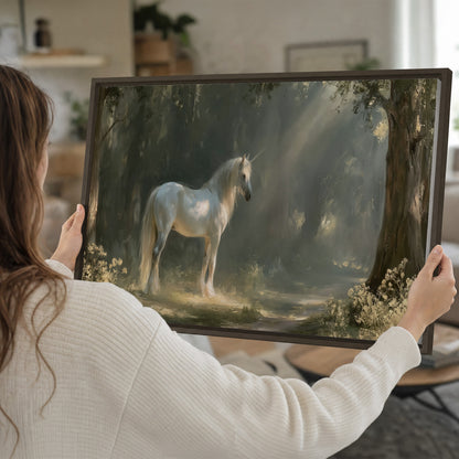 A white unicorn standing beneath arching trees in a softly lit woodland single framed canvas wall art held in hands