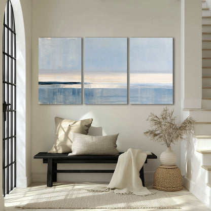 A minimal blue seascape with a pale horizon and soft vertical reflections 3 piece canvas wall art displayed in a entryway