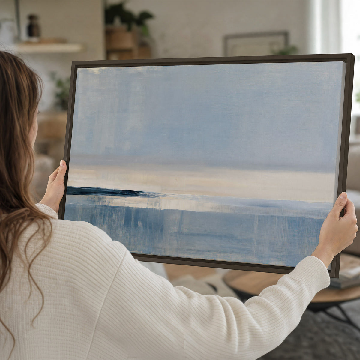 A minimal blue seascape with a pale horizon and soft vertical reflections single framed canvas wall art held in hands