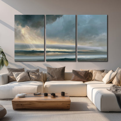 Dramatic blue-gray storm clouds breaking over a pale strip of ocean light 3 piece canvas wall art displayed in a living room