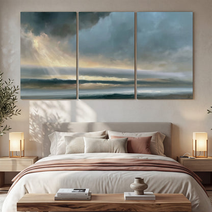 Dramatic blue-gray storm clouds breaking over a pale strip of ocean light 3 piece canvas wall art displayed in a bedroom